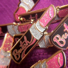 Load image into Gallery viewer, A collection of gold enamel pins of a open black tube of pink glittery lipstick with "Butch" in pink glitter is written on the side of the tube on top of fuchsia fabric