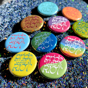 A collection of buttons in varying colors and patterns (solid blue, orange, teal, and yellow, metallic gold and copper, half pink and yellow, half blue and green, and rainbow stripes) that all say "pride was a RIOT" across the whole button. 
