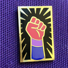 Load image into Gallery viewer, Enamel pin of a raised fist colored like the bisexual flag with gold outlining against a black background with a gold sunburst sitting on top of purple fabric. 