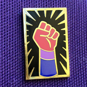 Enamel pin of a raised fist colored like the bisexual flag with gold outlining against a black background with a gold sunburst sitting on top of purple fabric. 