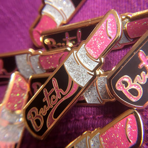 A collection of gold enamel pins of a open black tube of pink glittery lipstick with "Butch" in pink glitter is written on the side of the tube on top of fuchsia fabric