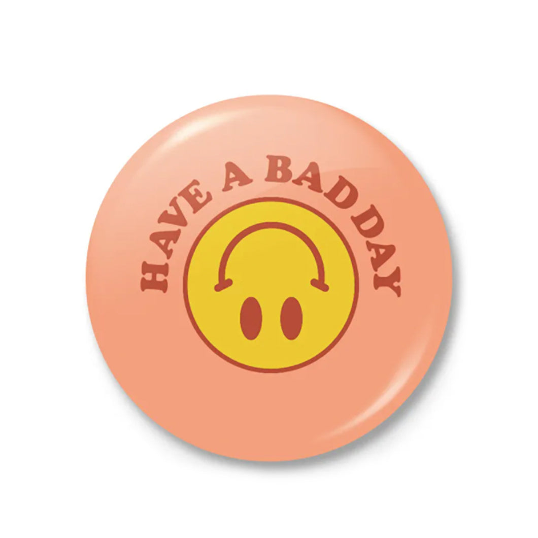 This larger, 2.5-inch button features a peach background, dark orange text saying “Have A Bad Day” in a half circle over an upside down smiley face, and has a latch mechanism backing. 