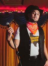 Load image into Gallery viewer, A picture of a model holding a microphone on a stand wearing the t-shirt, a black vest, red bandana, and cowboy hat on a stage.