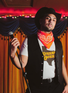 A picture of a model holding a microphone on a stand wearing the t-shirt, a black vest, red bandana, and cowboy hat on a stage.