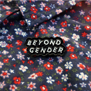 Enamel pin that reads "BEYOND GENDER" in white text angled diagonally upwards with a black border placed under the collar of a flower printed button-up shirt.