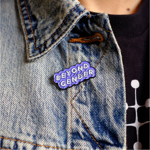 Enamel pin that reads "BEYOND GENDER" in white text angled diagonally upwards with a light purple border fastened underneath the collar of a jean jacket.  