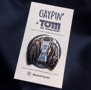 A picture of an enamel pin of a leather jacket with a white shirt with art of a shirtless man in black jeans and a conductor hat by Tom of Finland underneath on a white card with black text that reads "Gaypin' + Tom of Finland" along the top.