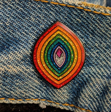 Load image into Gallery viewer, Enamel pin of a pointed oval with sparkly rainbow sections in a vagina shape placed on a piece of denim.