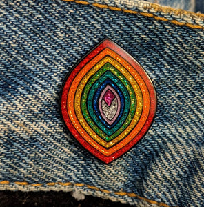 Enamel pin of a pointed oval with sparkly rainbow sections in a vagina shape placed on a piece of denim.