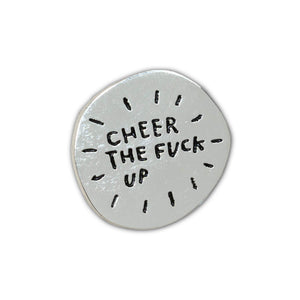 A silver enamel pin in the shape of an imperfect circle that reads "Cheer The Fuck Up" with a simple black starburst lines around the text.