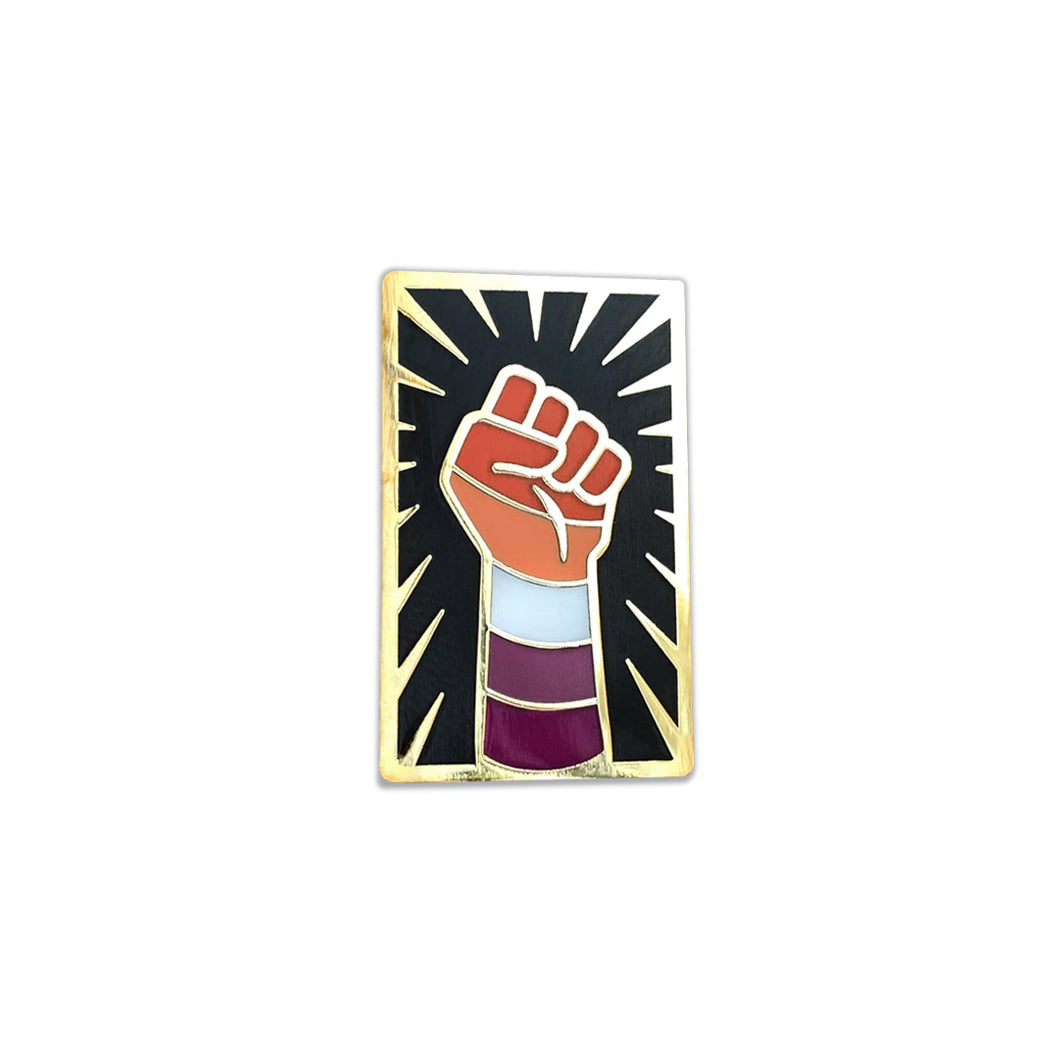 Enamel pin of a raised fist colored like the lesbian flag with gold outlining against a black background with a gold sunburst.