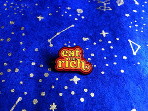 An enamel pin that reads "Eat the Rich" in 70's style yellow font outlined by orange and then red sitting on top of a blue background with constellations on it. 