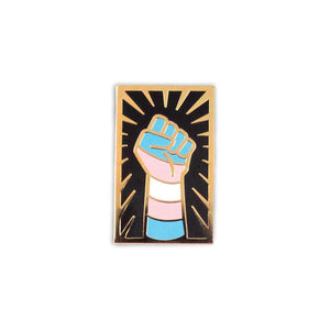 Enamel pin of a raised fist colored like the transgender flag with gold outlining against a black background with a gold sunburst.
